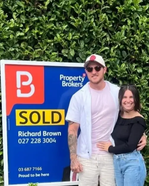 Couple in front of a hedge beside their Sold sign